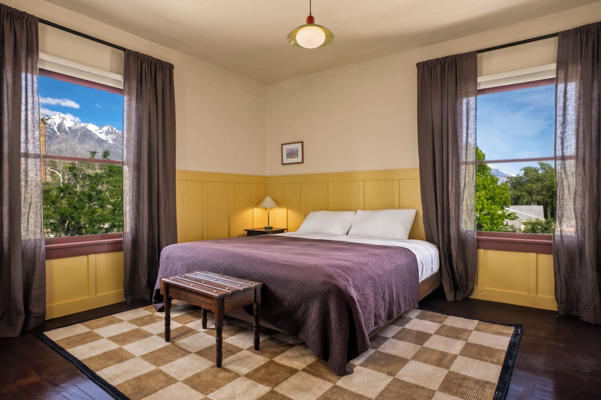 A bedroom with a bed, a nightstand, a lamp, a window with curtains, and a view of mountains.