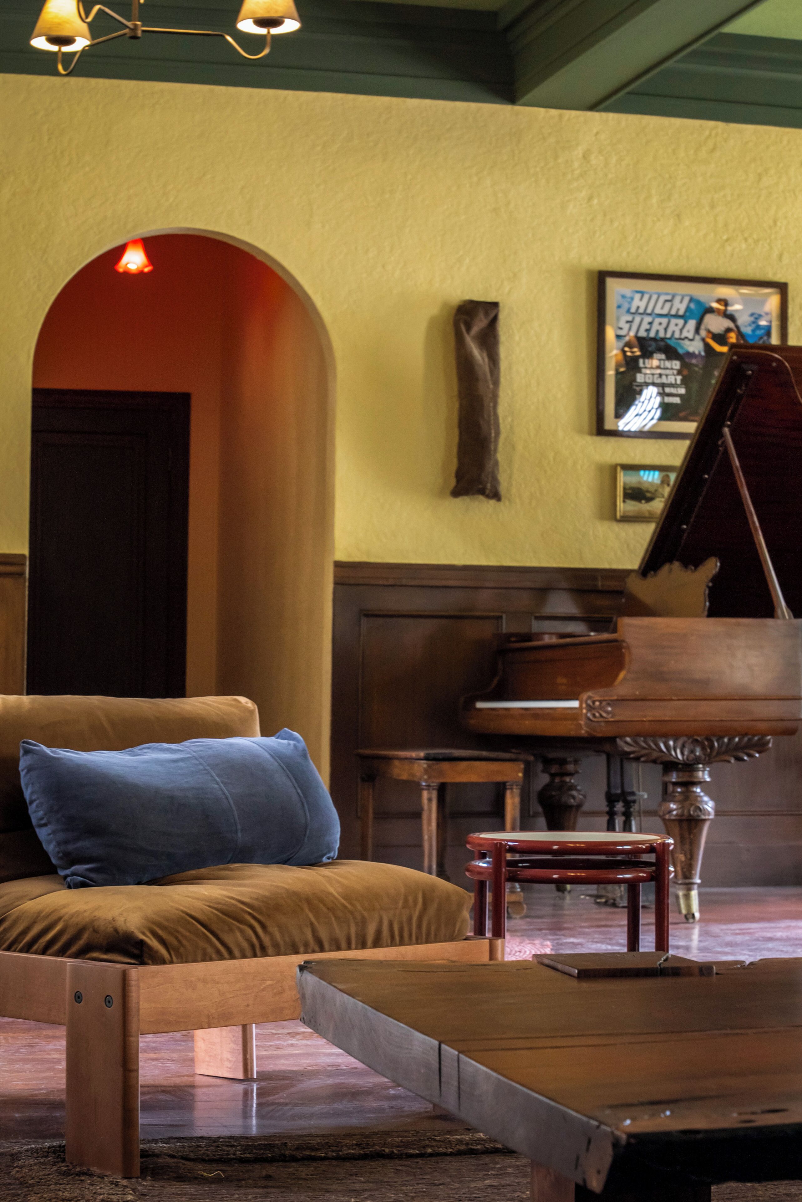 A room with a piano, a sofa with a blue pillow, a wooden bench, and a framed poster on the wall.