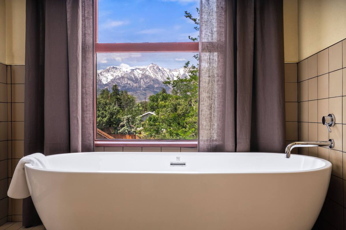 A bathroom with a bathtub, a window with curtains, and a view of mountains.