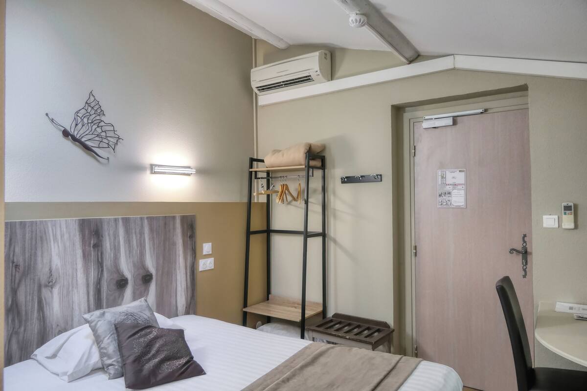 A hotel room with a bunk bed, a wall-mounted shelf, a bed with pillows, a bedside table, a chair, and an air conditioning unit.