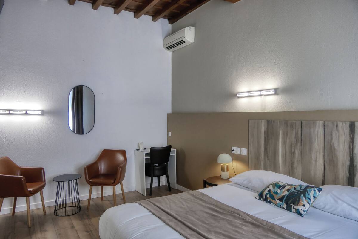 A hotel room with a bed, a desk with a chair, a round table, and a wall-mounted air conditioner.