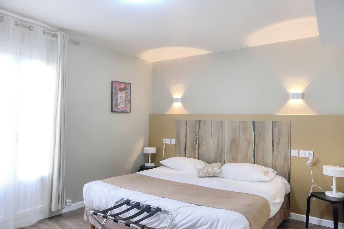 A hotel room with a bed, bedside lamps, a headboard, a nightstand, a wall-mounted light, and a window with curtains.