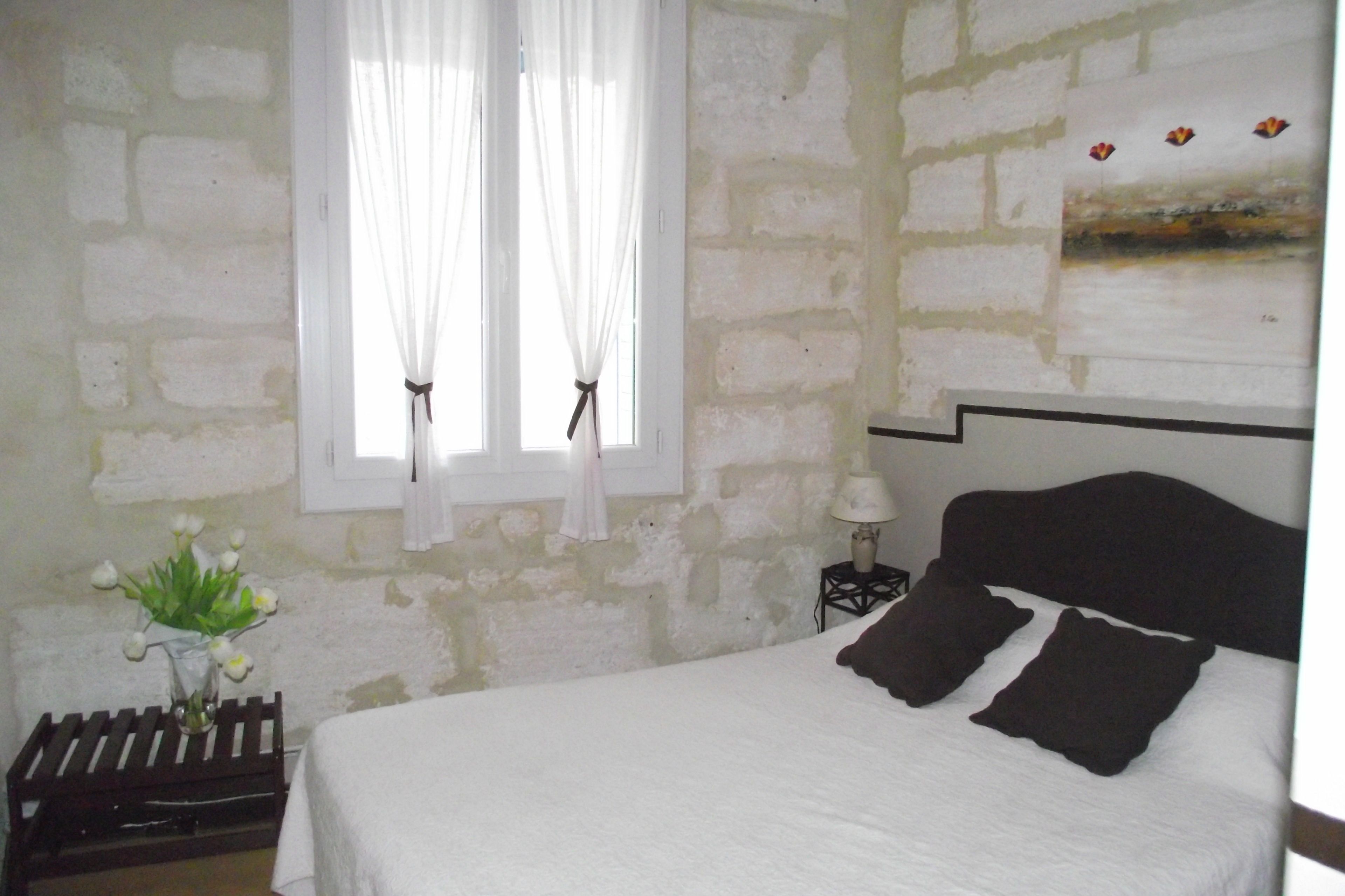 A bedroom with a bed, a nightstand, a lamp, a window with curtains, and a wall with a painting.