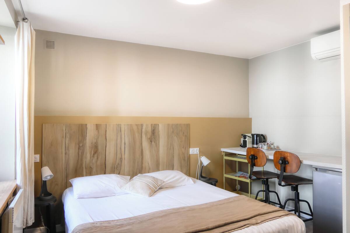 A hotel room with a bed, a desk with two chairs, a small table with a kettle, and a wall-mounted air conditioner.
