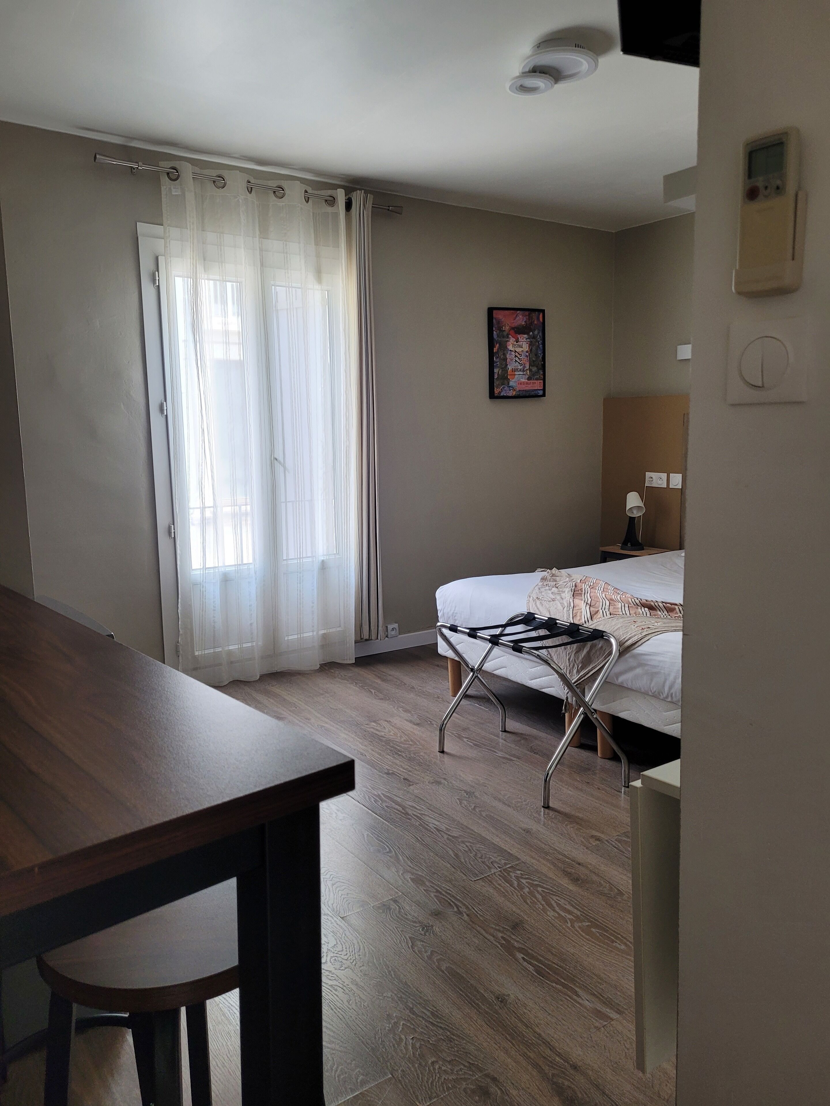 A bedroom with a bed, a desk, a chair, a window with curtains, a wall-mounted heater, and a poster on the wall.