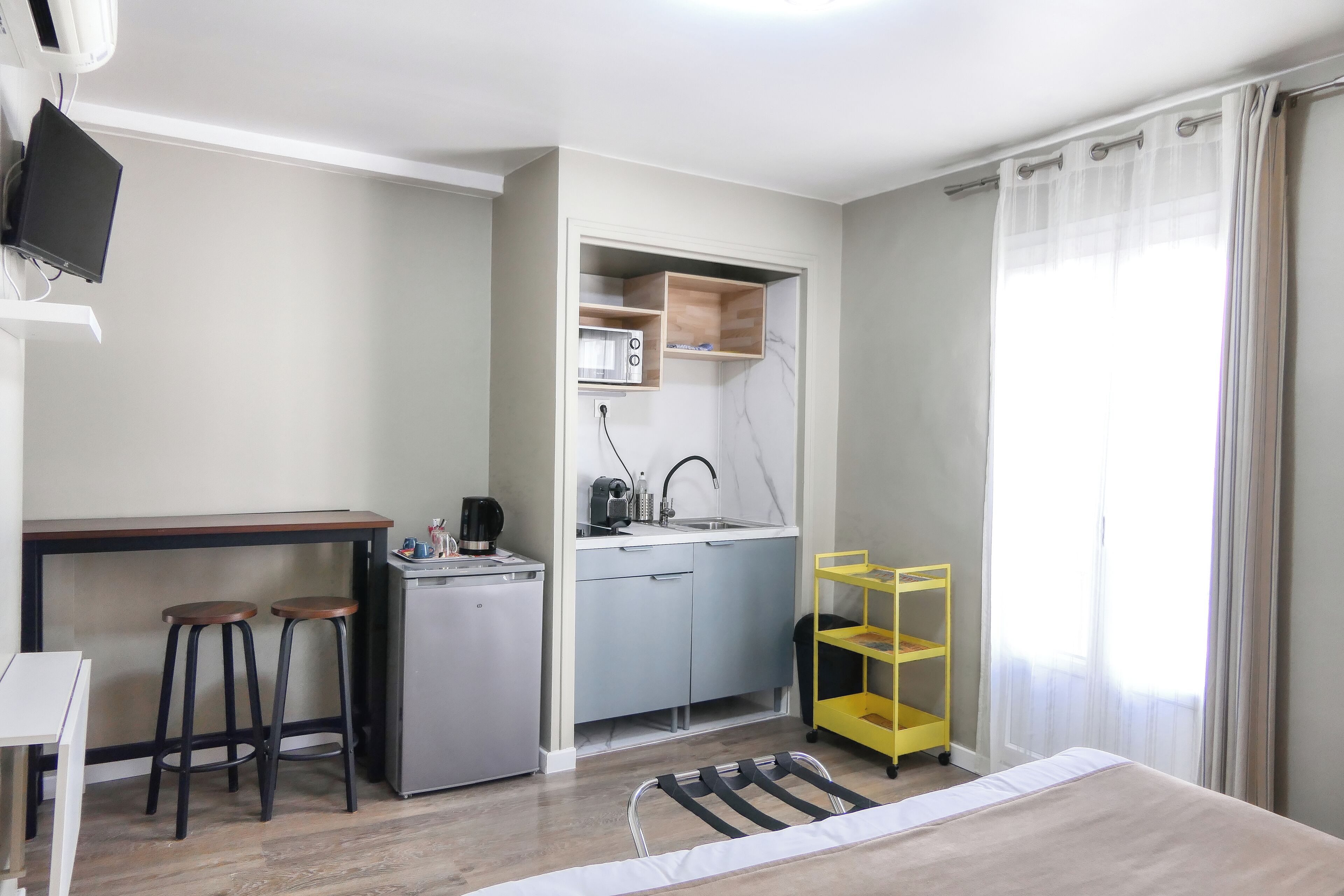 Een compacte kamer met een kitchenette, een bed, een bureau met twee krukken en een aan de muur bevestigde televisie.