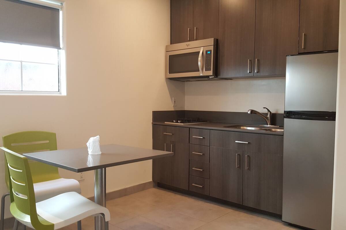 A compact kitchen with dark wood cabinets, a stainless steel refrigerator, a microwave, and a sink. There is a small table with a chair and a tissue box on it.