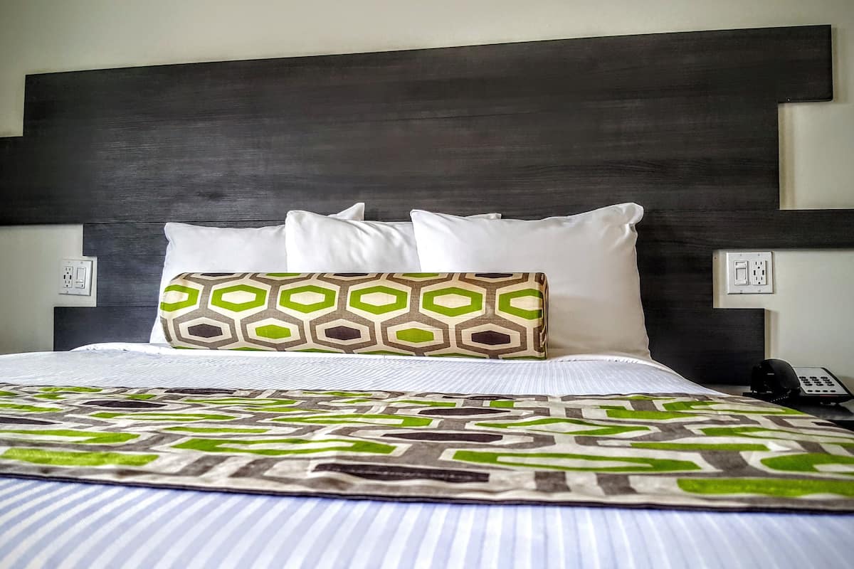 A hotel room with a large bed, a dark headboard, and a patterned throw blanket.