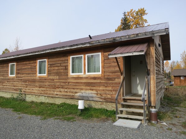 A wooden cabin with a metal roof, a front porch with steps, and a small front yard. - All Seasons Campground & Cabins (Ninilchik)