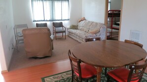 A living room with a wooden dining table, two chairs, a sofa, and a window with curtains. - All Seasons Campground & Cabins (Ninilchik)