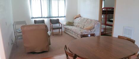 A living room with a wooden dining table, two chairs, a sofa, and a window with curtains.