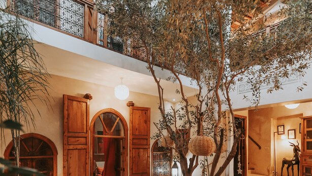 A spacious indoor area with a central wooden table, benches, and a large tree. There are wooden doors and a staircase leading to an upper level. - Dar Nour El Houda (Marrakech)