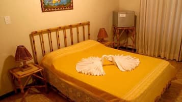 Quarto de hotel com cama, abajur, televisão em suporte e cortina.