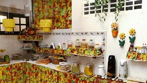 A kitchen with a colorful floral tablecloth, shelves with food items, and hanging plants. - Pousada Montreux (Mata de São João)