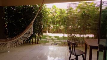 A patio area with a hammock, a wooden chair, and a table. There is a view of a garden with various plants and a pathway leading to a door.