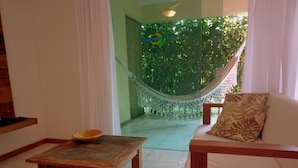 Una sala de estar con una mesa de centro de madera, un sofá blanco con un cojín estampado, una hamaca y vistas a la vegetación a través de un ventanal.