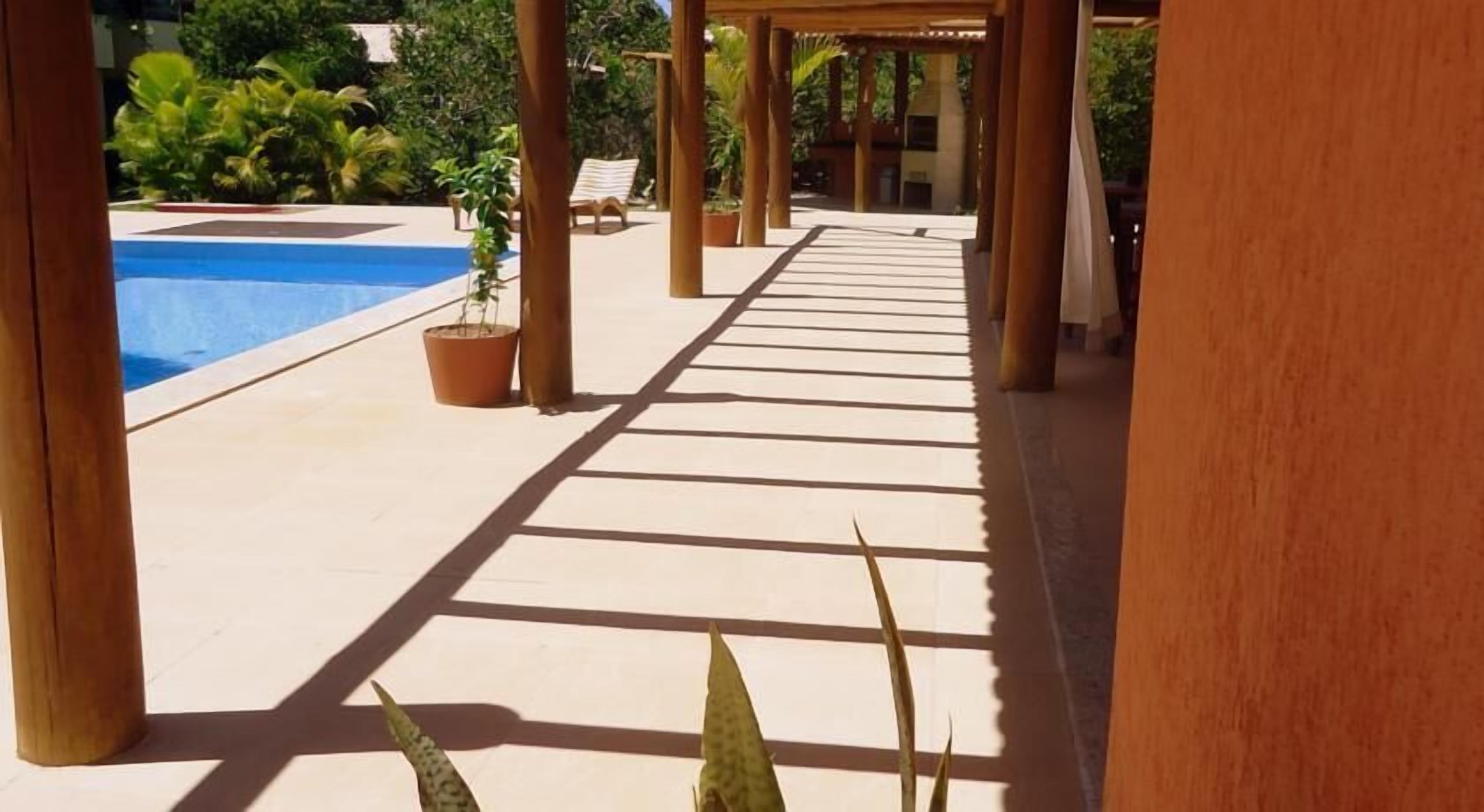 Un paseo techado con piscina a un lado, plantas en macetas y cielo despejado.