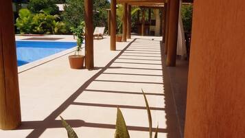 Un paseo techado con piscina a un lado, plantas en macetas y cielo despejado.
