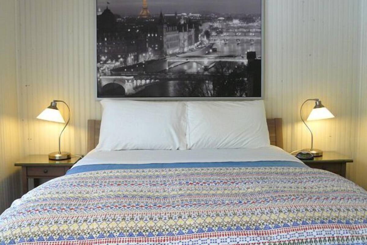 A hotel room with a bed, two bedside lamps, a large cityscape photograph on the wall, and a patterned bedspread.