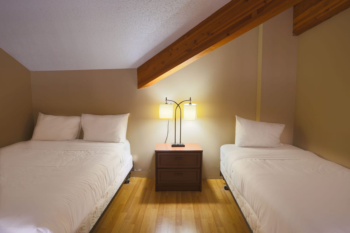 Un dormitorio con dos camas, una mesita de noche con lámpara, vigas de madera en el techo y una pared lisa.