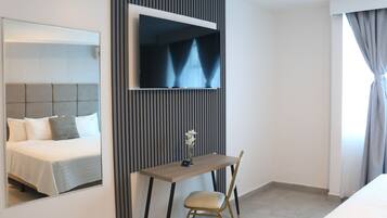 Habitación de hotel con cama, escritorio, silla, televisor montado en la pared y ventana con cortinas.