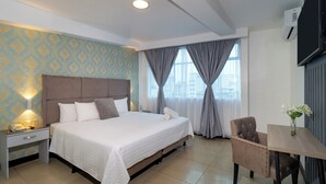 Een hotelkamer met een groot bed, een nachtkastje met een lamp, een stoel, een tafeltje met een vaas en een raam met gordijnen.