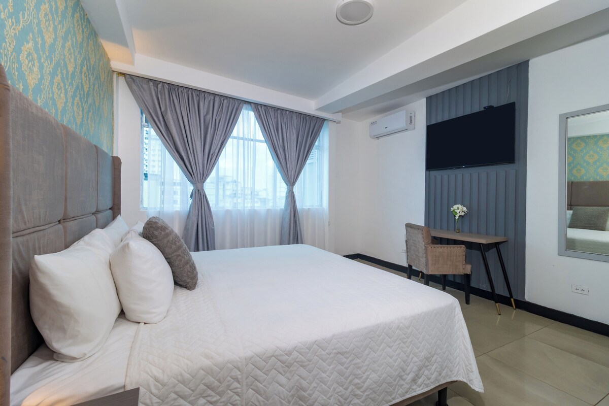 A hotel room with a bed, a desk, a chair, a TV, and an air conditioning unit.