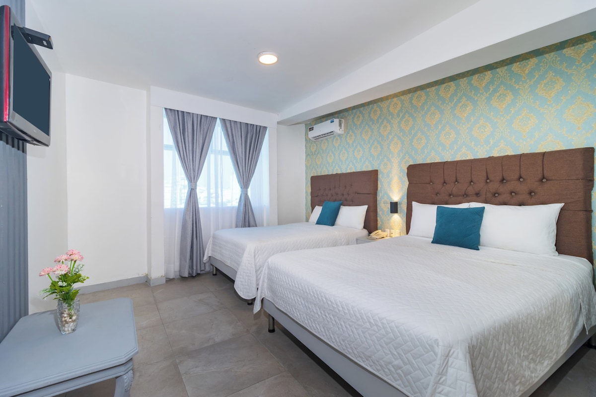 A hotel room with two beds, a TV, a vase with flowers, and a window with curtains.