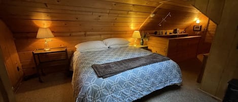 Un dormitorio acogedor en el ático, con una cama, dos lámparas de noche, una mesita de noche y un aparador de madera.