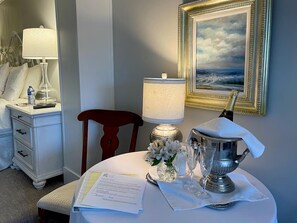 A neatly arranged hotel room with a bed, a nightstand with a lamp, a chair, a table with a vase of flowers, a framed painting, and a bottle of champagne. - A Vista Villa Couples Retreat (Kelowna)