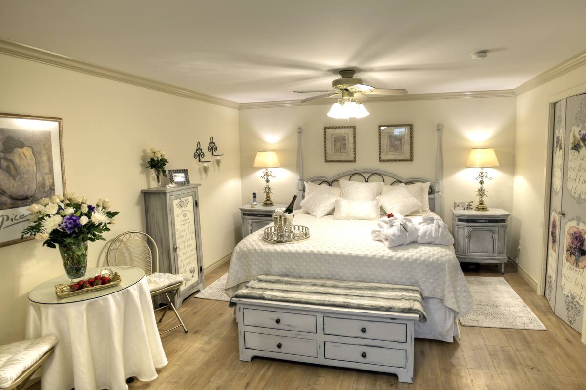 A bedroom with a bed, bedside tables, a chair, a small table with a vase of flowers, and a ceiling fan.