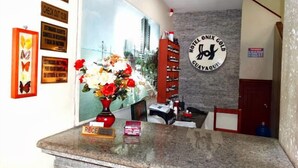Zona de recepción de un hotel con mostrador, un jarrón de flores, un cartel en la pared con el logotipo del hotel y un ventanal con vistas a la ciudad.