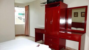 Una habitación de hotel con una cama, un armario rojo, un televisor encima del armario y una ventana con cortinas.