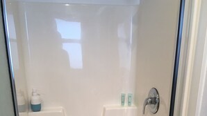 A clear shower area with a drain, a white tiled wall, and a door with a silver handle. - All Seasons Bed & Breakfast (Kelowna)
