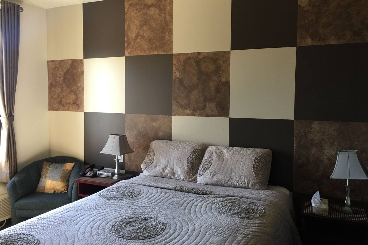 Ein Hotelzimmer mit Schachbrettmuster an der Wand, einem Bett mit gesteppter Bettdecke, einem Sessel und zwei Nachttischen mit Lampen und einer Papiertüte.