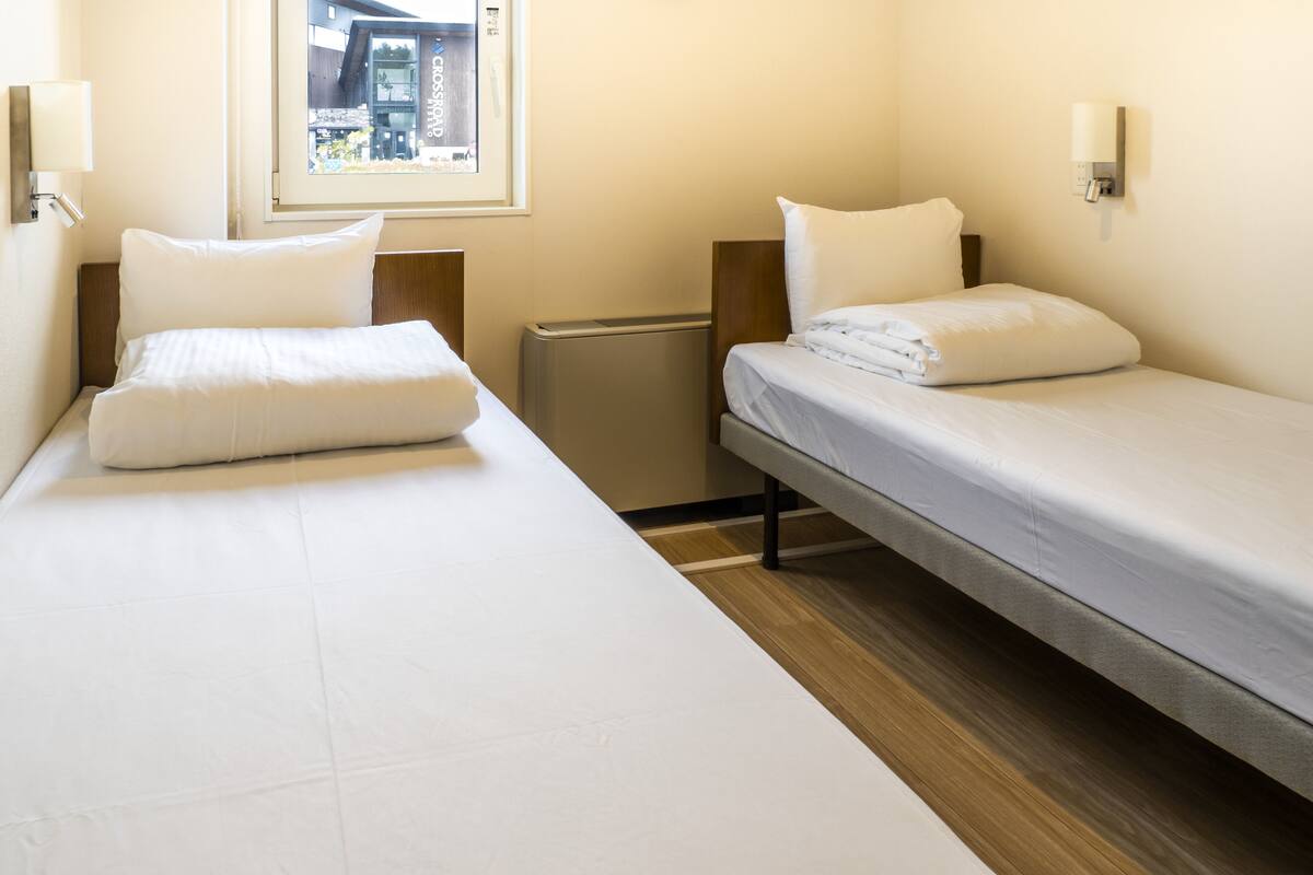 A hotel room with two single beds, a small heater, and a window with a view of the city.