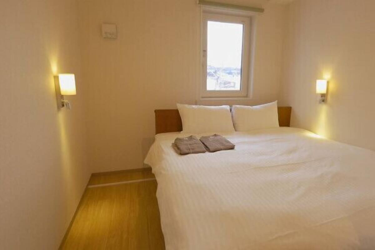 A neatly made bed with pillows and a blanket, a window with a view, and two wall-mounted lights.