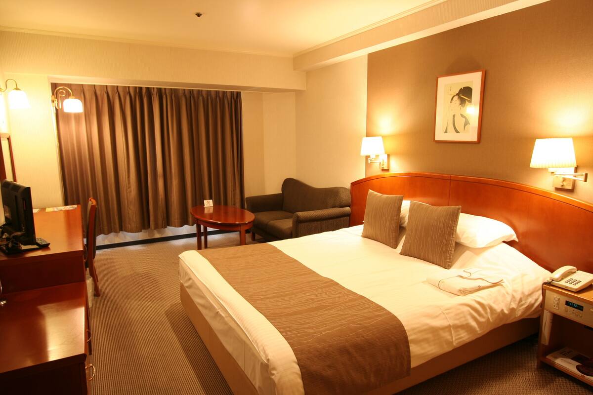 Ein Hotelzimmer mit einem großen Bett, einem Schreibtisch, einem Sessel, einem kleinen Tisch und einem Nachttisch mit Telefon.