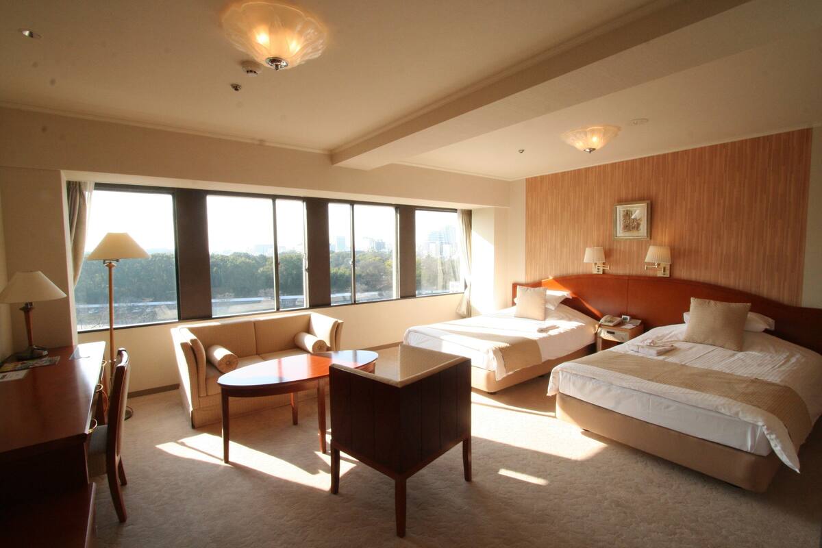 Ein Hotelzimmer mit einem großen Bett, zwei Nachttischen, einem Schreibtisch mit Stuhl, einem Sofa und Blick auf die Stadt durch große Fenster.