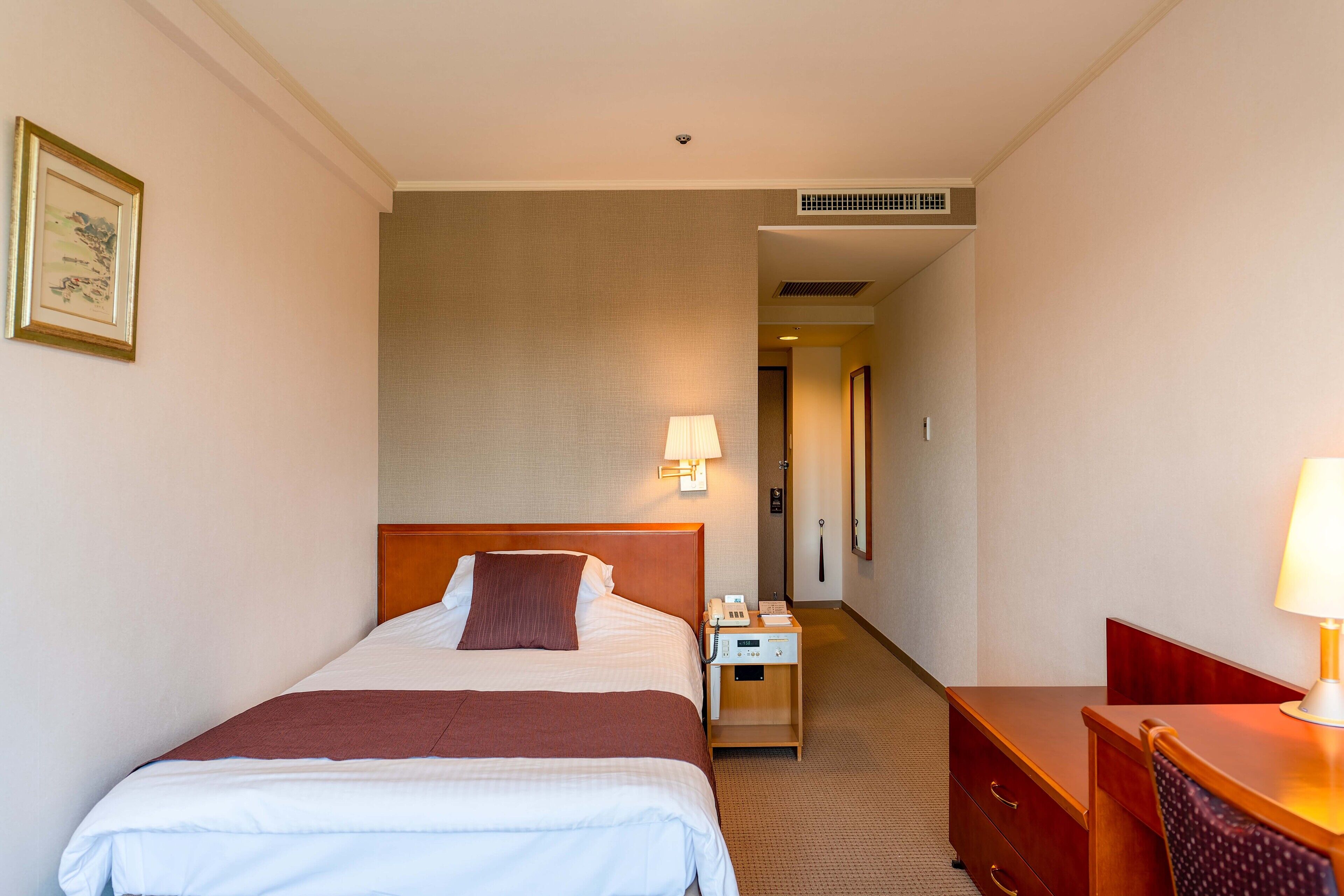 A hotel room with a large bed, a bedside table with a lamp, a desk with a chair, and a framed picture on the wall.