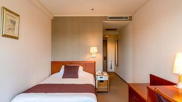 A hotel room with a large bed, a bedside table with a lamp, a desk with a chair, and a framed picture on the wall.