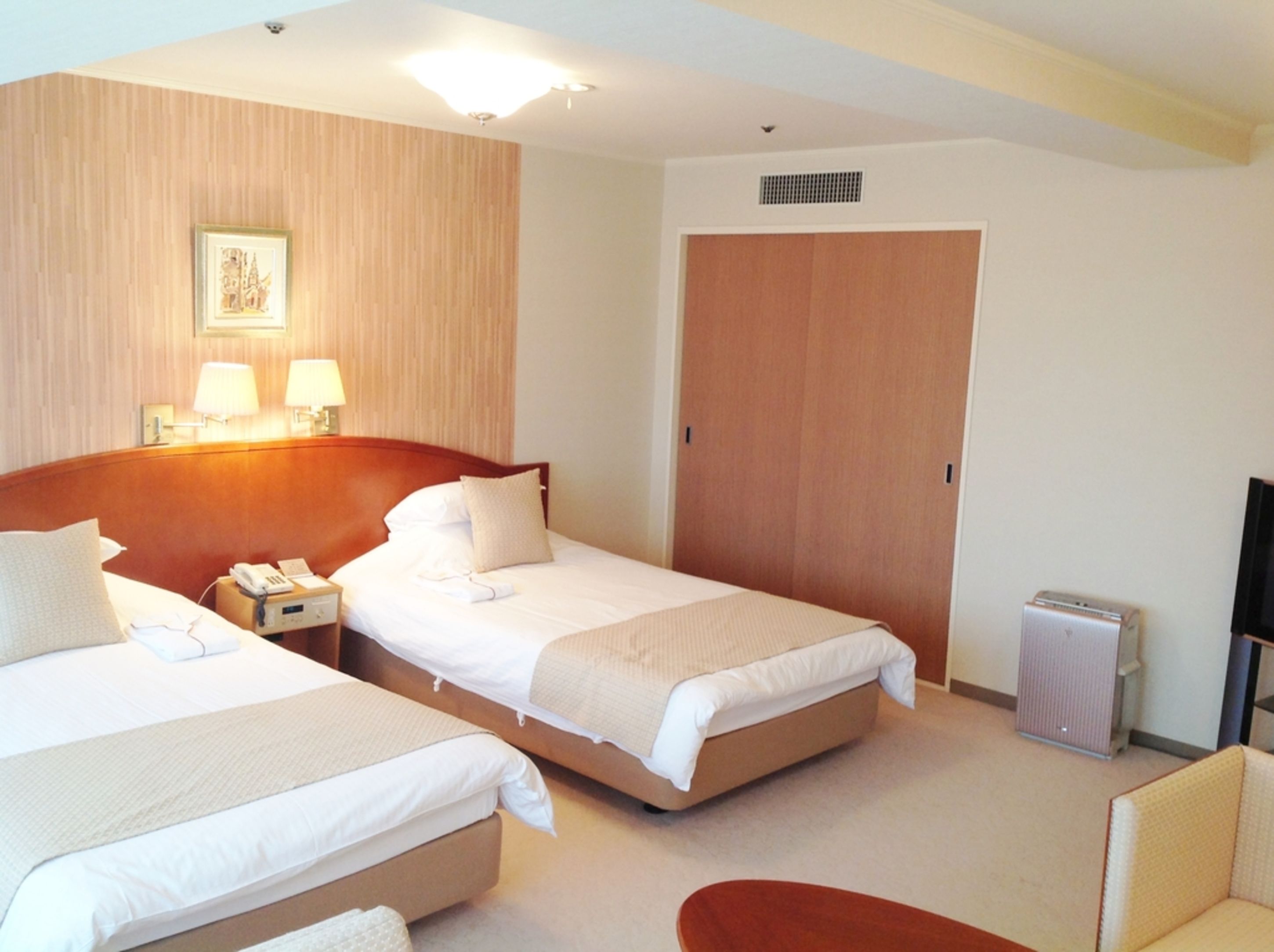 A hotel room with two beds, a nightstand with a phone, a lamp, a heater, and a TV.
