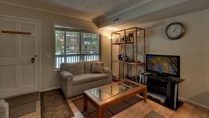 A living room with a sofa, a glass coffee table, a TV on a stand, and a bookshelf. - 1200 Wildwood Unit 16 (South Lake Tahoe)