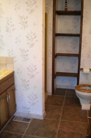 A bathroom with floral wallpaper, a wooden vanity, a toilet with a wooden seat, and a shelf unit.
