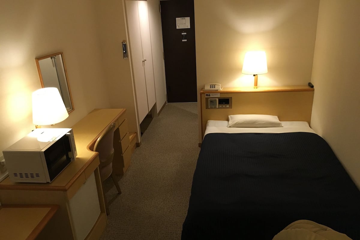 A hotel room with a bed, a desk with a lamp, a microwave, a telephone, and a door with a sign.