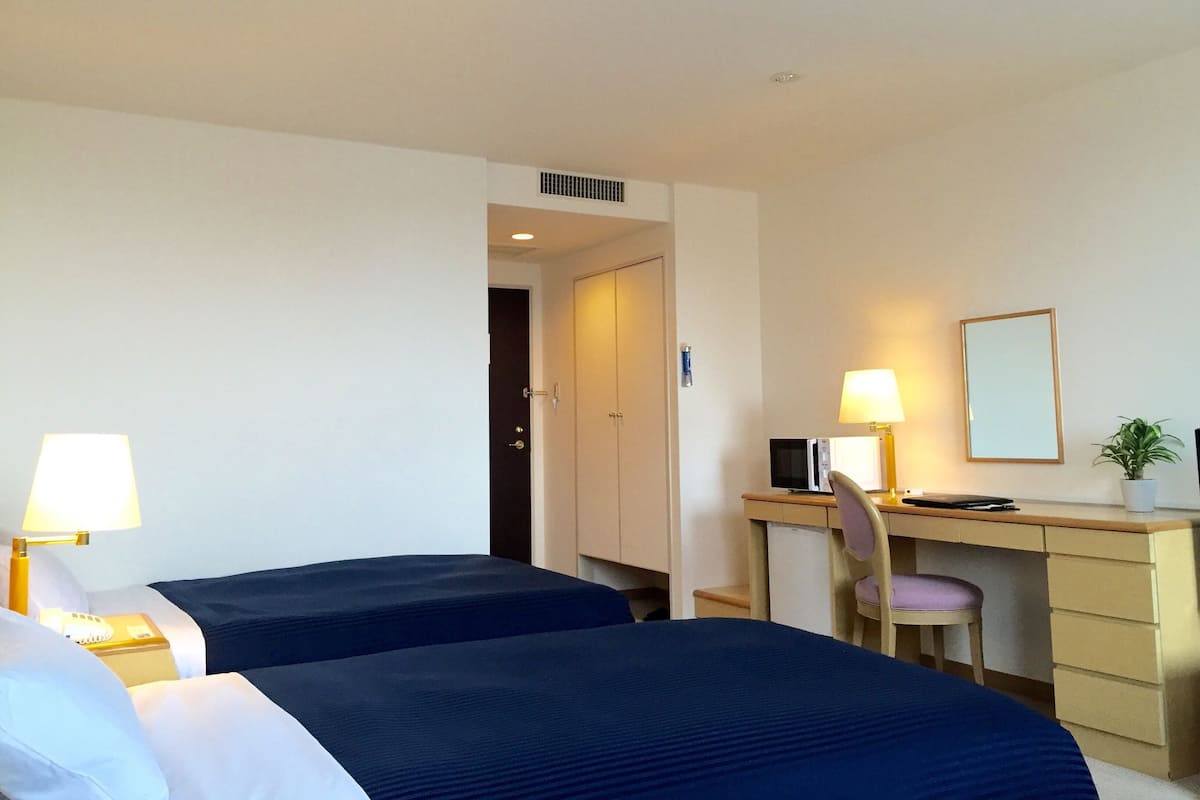 A hotel room with two beds, a desk, a chair, a lamp, a mirror, and a potted plant.