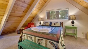 Un dormitorio acogedor en el ático, con techo inclinado, una cama con colcha estampada, dos lámparas de noche y una ventana con vista a árboles.
