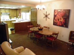 A dining area with a wooden table and chairs, a kitchen in the background, and a painting on the wall.