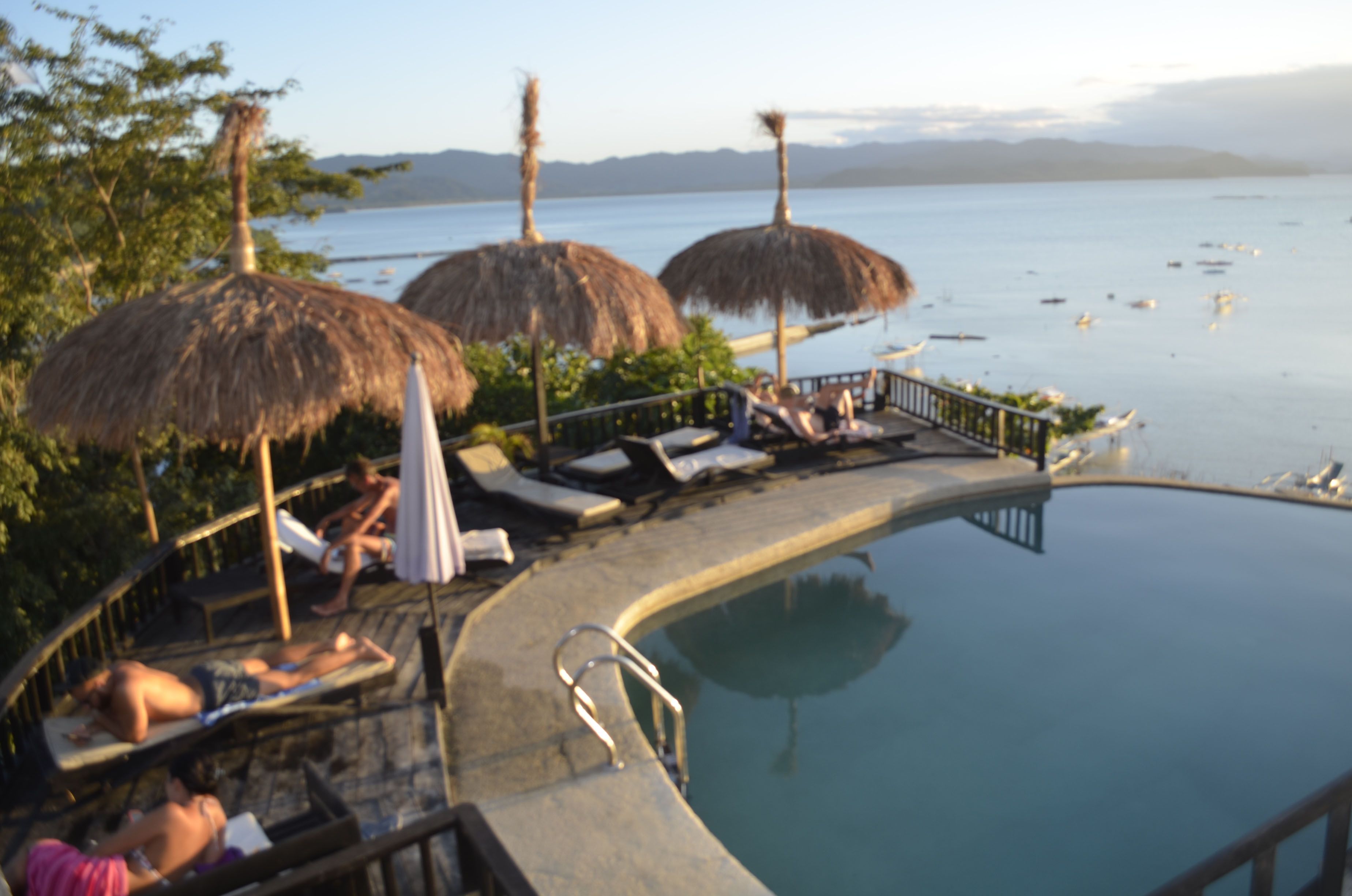 A poolside area with lounge chairs and thatched umbrellas, overlooking a calm body of water.