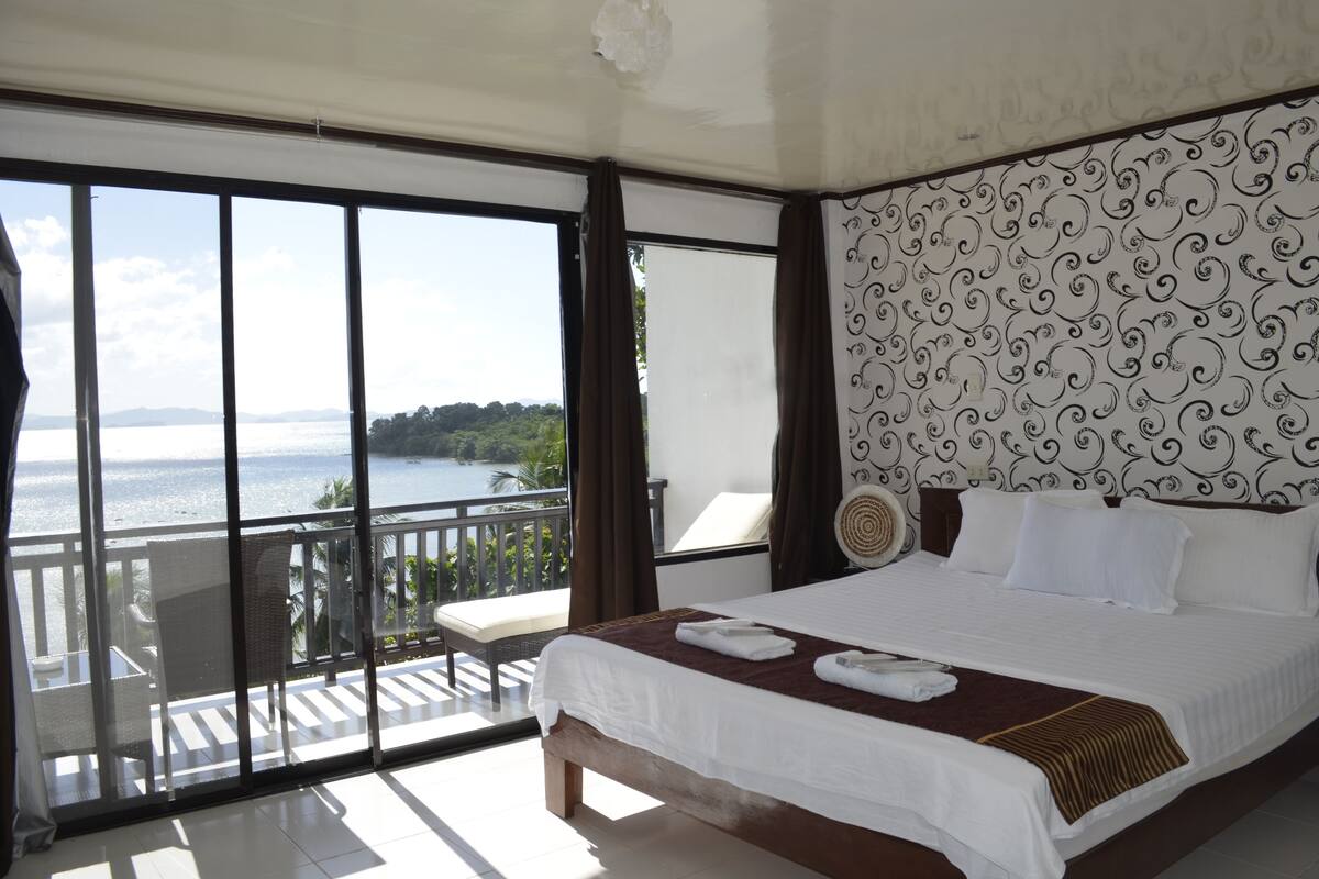 A hotel room with a large bed, a balcony with a view of the sea, and a patterned wall.